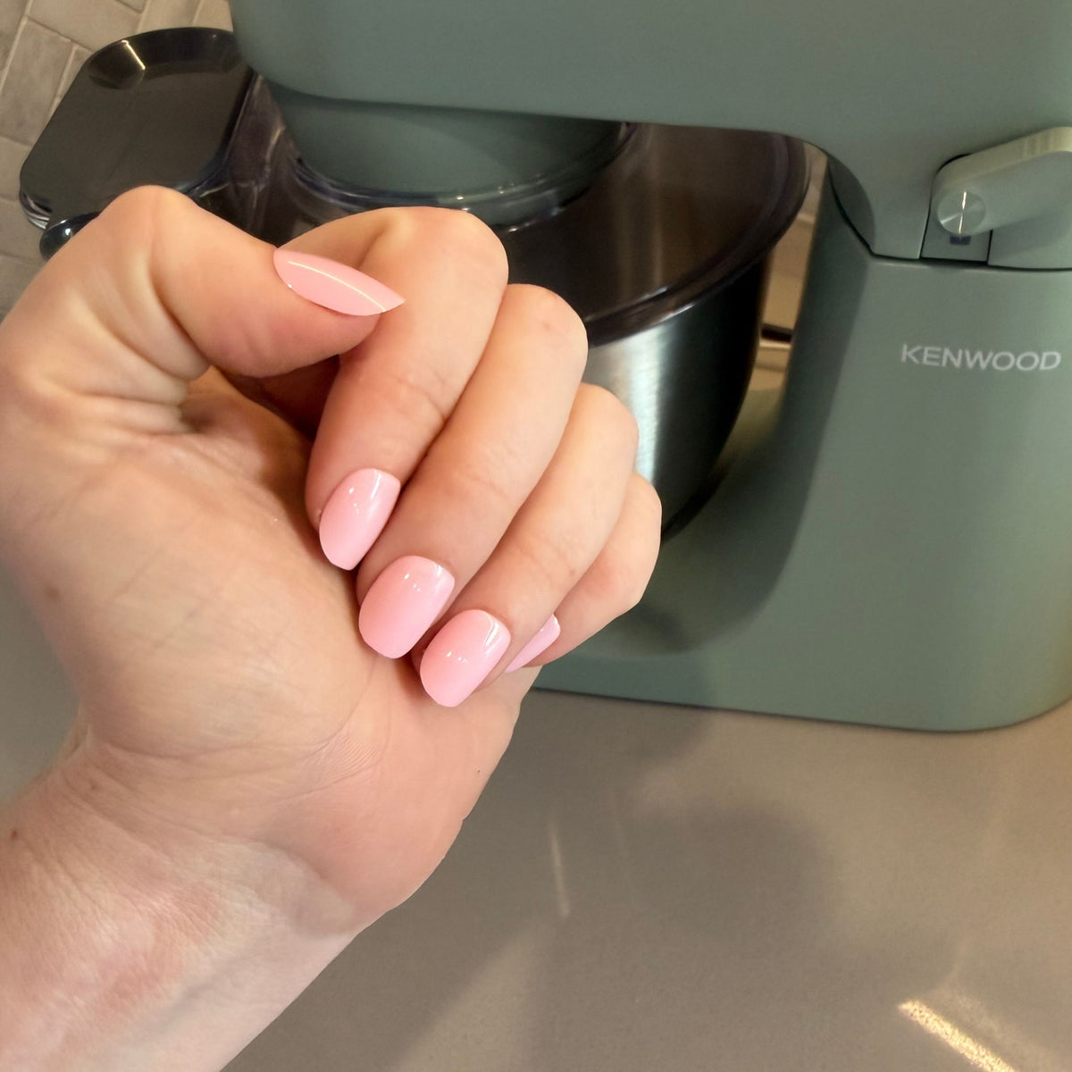 A close-up of a hand with a soft light pink manicure, positioned in front of a sage green Kenwood stand mixer on a countertop.