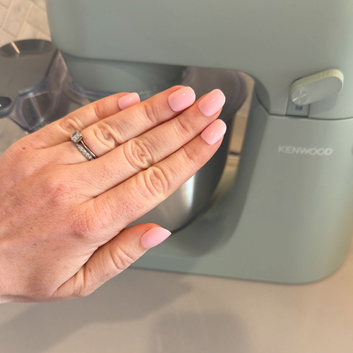 A close-up of a hand with neatly manicured light pink nails resting in front of a Kenwood countertop appliance.