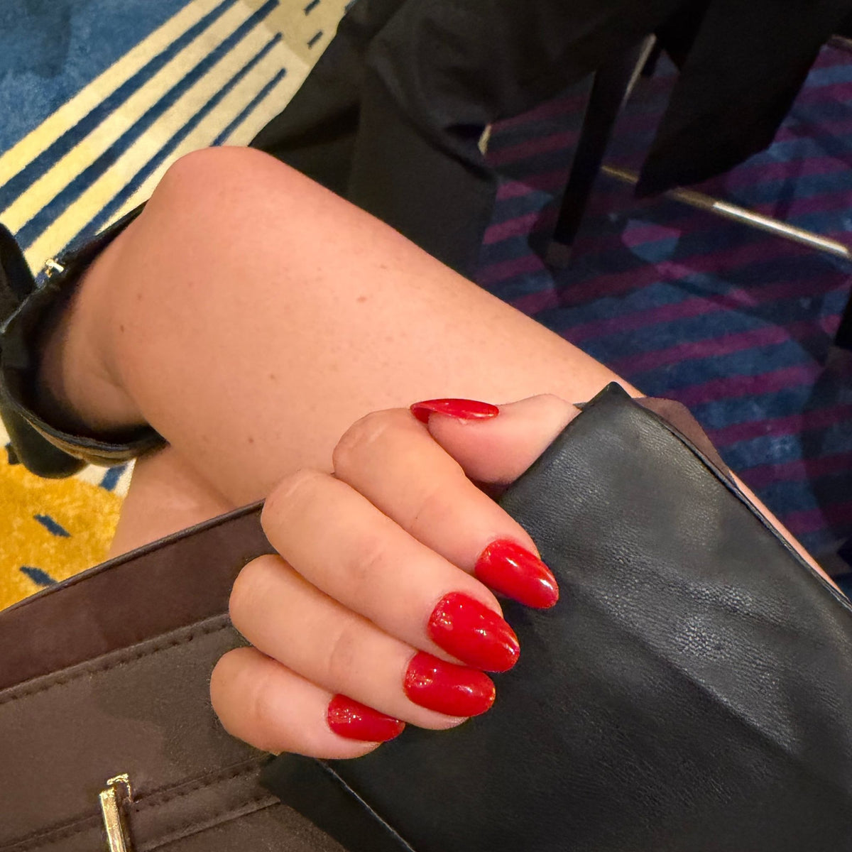 A close-up of a hand with glossy red manicured nails resting on a black leather surface, with a person's leg visible in the background.