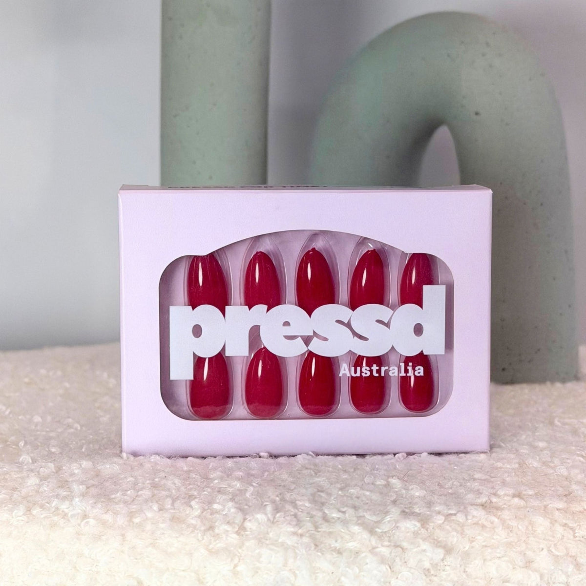 A box of red press-on nails with the brand name 'pressd' and 'Australia' printed on the front, set on a textured white surface with a soft-focus background.
