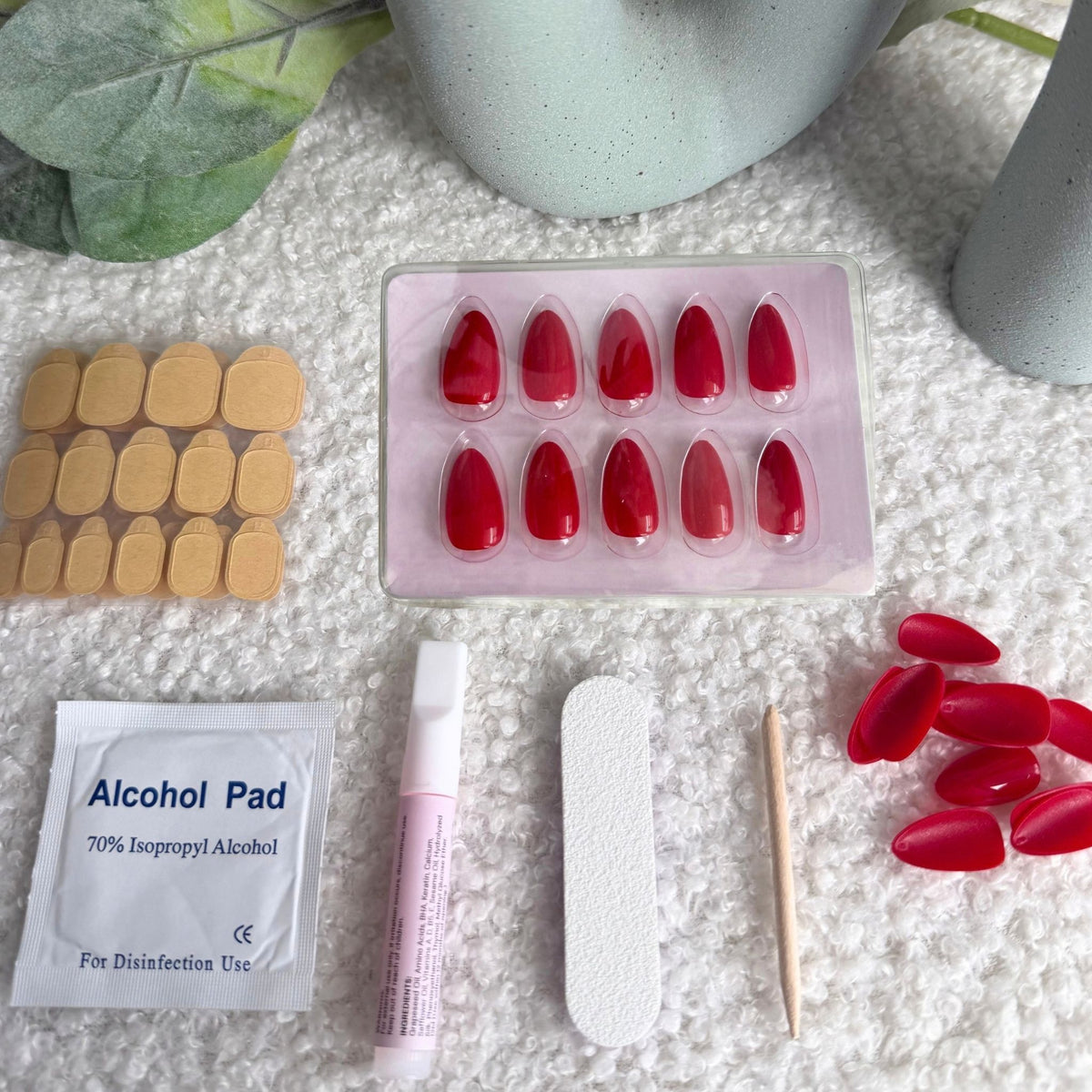 A flat-lay of press-on nail kit components on a white textured surface, including a tray of red false nails, a sheet of beige adhesive stickers, a packet of alcohol pads, a pink nail file, a wooden nail tool, and a small tube.