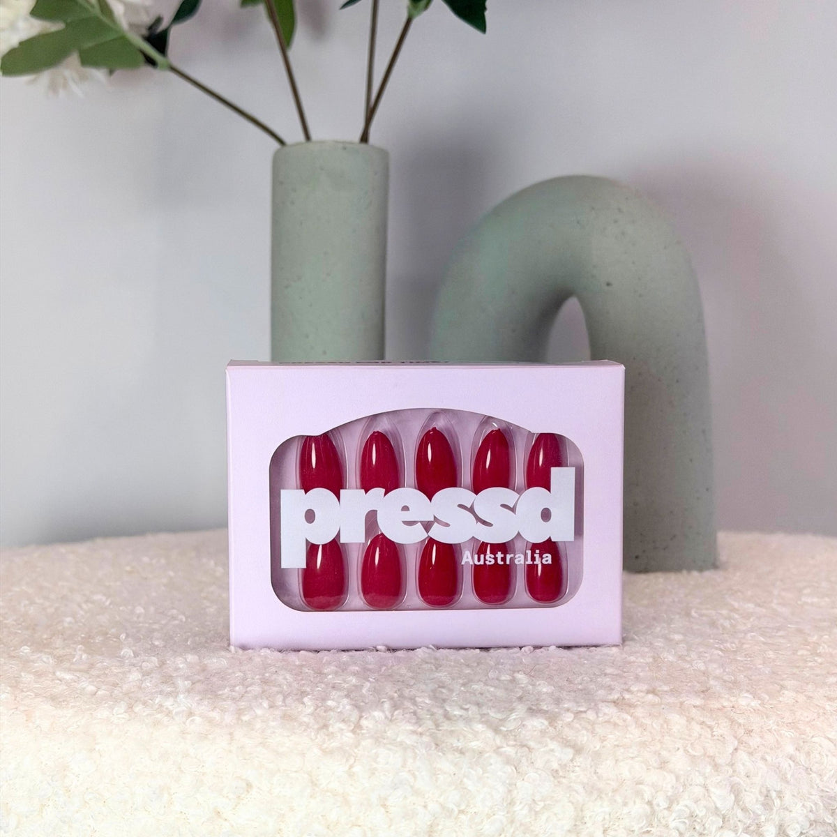 A set of red press-on nails in a lilac box with the brand name 'pressd australia' visible on the front, styled on a cream textured surface with a green ceramic vase in the background.